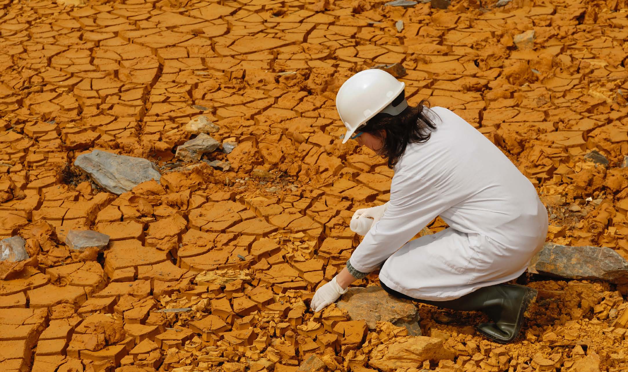 Female climate scientist collecting soil samples in a dry, arrid desert with cracked orange dirt.