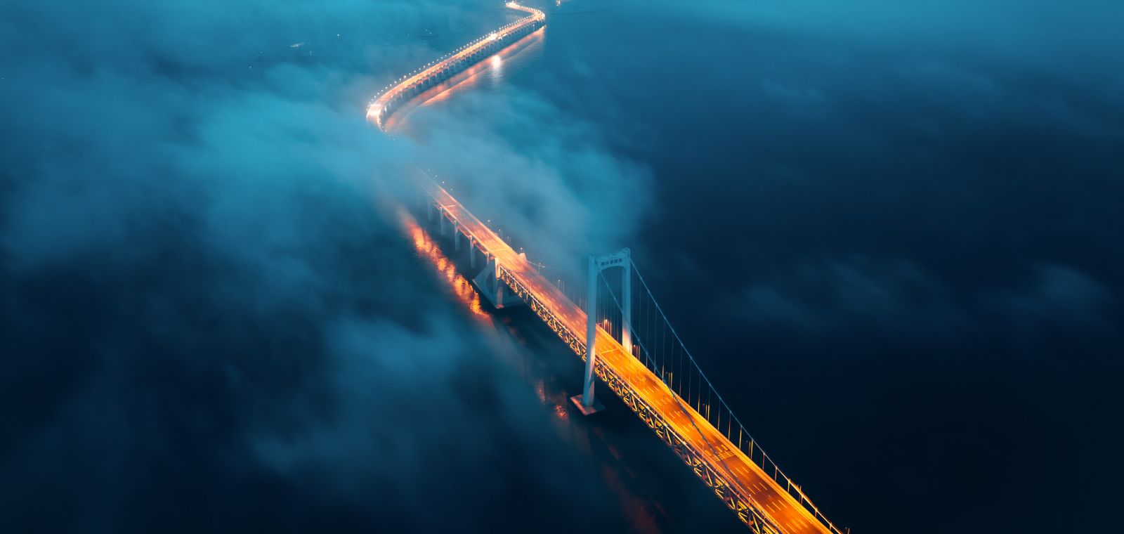 A long, illuminated bridge snakes through the evening mist toward a distant location.